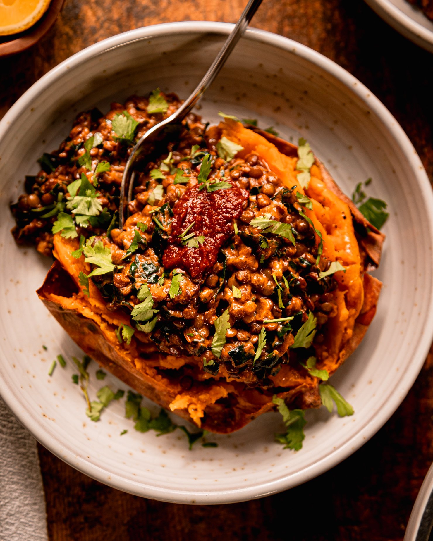 An overhead shot shows a baked sweet potato in a bowl that's stuffed with a creamy harissa coconut lentils and kale mixture. The dish is garnished with chopped cilantro and a dab of harissa.