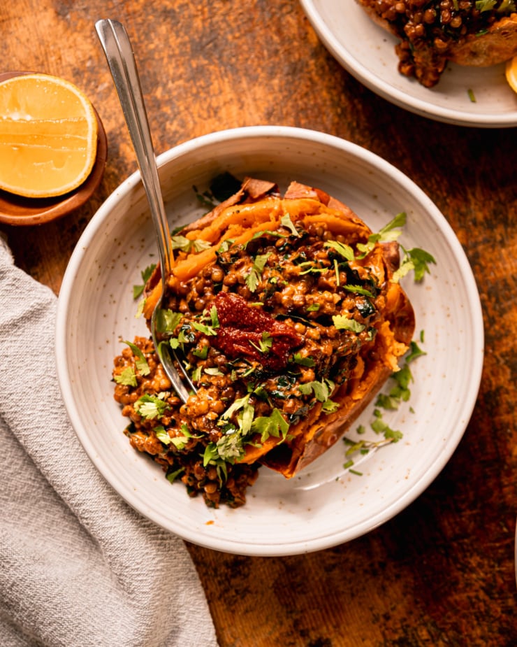 An overhead shot shows a baked sweet potato in a bowl that's stuffed with a creamy harissa coconut lentils and kale mixture. The dish is garnished with chopped cilantro and a dab of harissa. A lemon wedge is seen nearby.