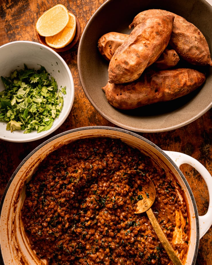 An overhead shot shows a lentil stew mixture in a pot, a bowl of whole roasted sweet potatoes, a bowl of chopped cilantro, and a couple big lemon wedges.