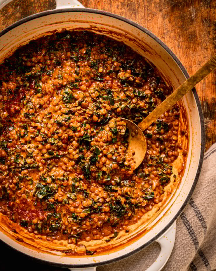 An overhead shot shows a braiser pot filled with harissa coconut lentils with kale. A ladle is sticking out of the pot.