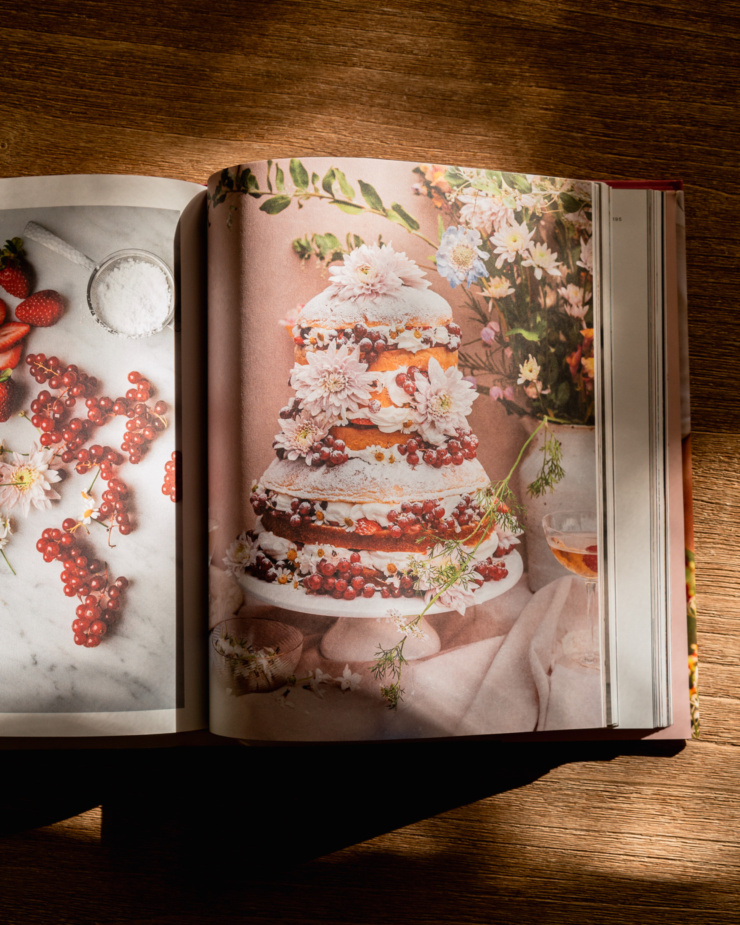 An overhead shot shows a cookbook open to a beautiful photo of a multi layer cake decorated with berries and flowers.