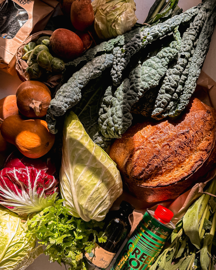 An overhead shot shows a variety of produce, a load of sourdough bread, olive oil, a bag of lentils, and shoyu.