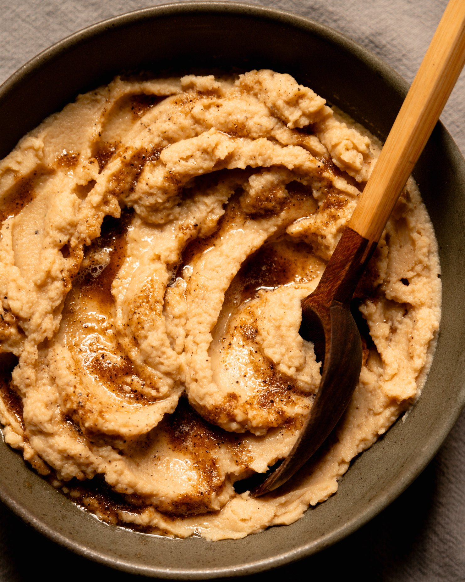 An overhead shot shows a bowl of mashed cauliflower with swoops of vegan Worcestershire black pepper butter on top. A serving spoon is sticking out of the mash.