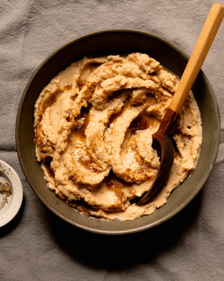An overhead shot shows a bowl of mashed cauliflower with swoops of vegan Worcestershire black pepper butter on top. A serving spoon is sticking out of the mash.