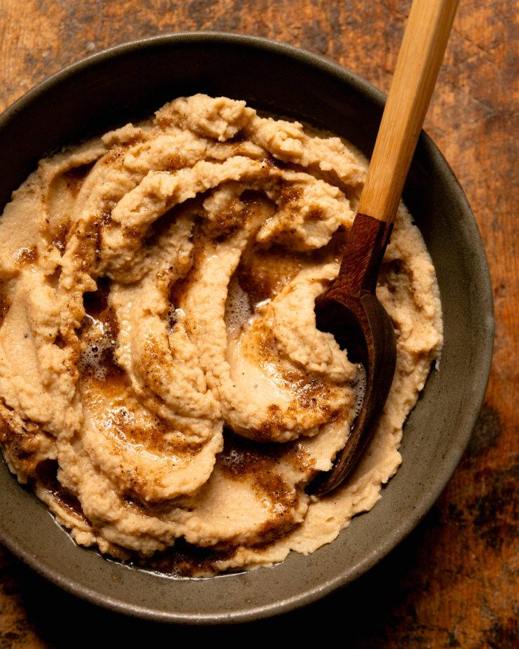 An overhead shot shows a bowl of mashed cauliflower with swoops of vegan Worcestershire black pepper butter on top. A serving spoon is sticking out of the mash.