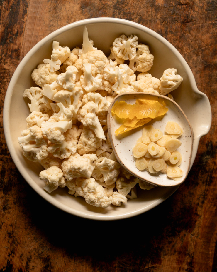 An overhead shot shows a large bowl filled with raw cauliflower florets. A smaller plate is perched on top that contains sliced garlic cloves and strips of lemon peel.