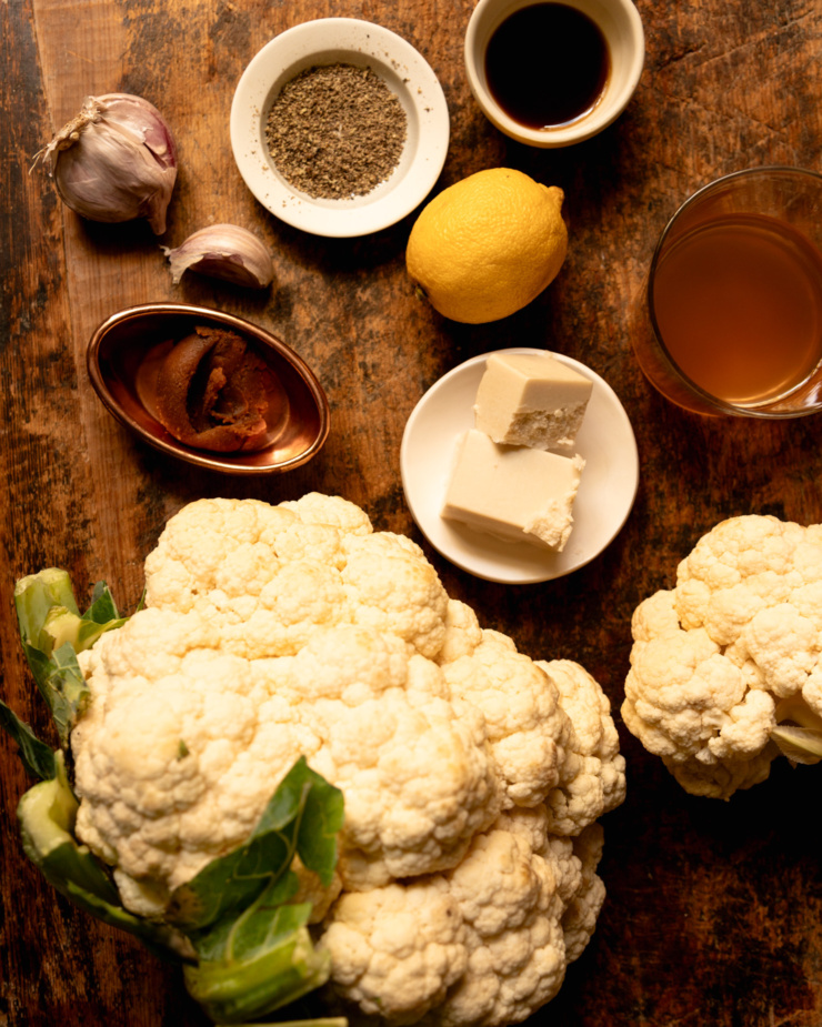 An overhead shot shows the following ingredients on a rough wood board: black pepper, vegan Worcestershire sauce, a lemon, vegan butter, vegetable stock, cauliflower, miso, and garlic.
