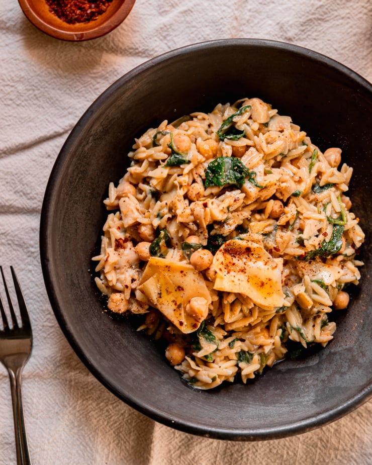 An overhead shot shows a single serving of a vegan spinach artichoke orzo dish. The bowl is wide and shallot. A bowl of ground chilies is nearby for topping.