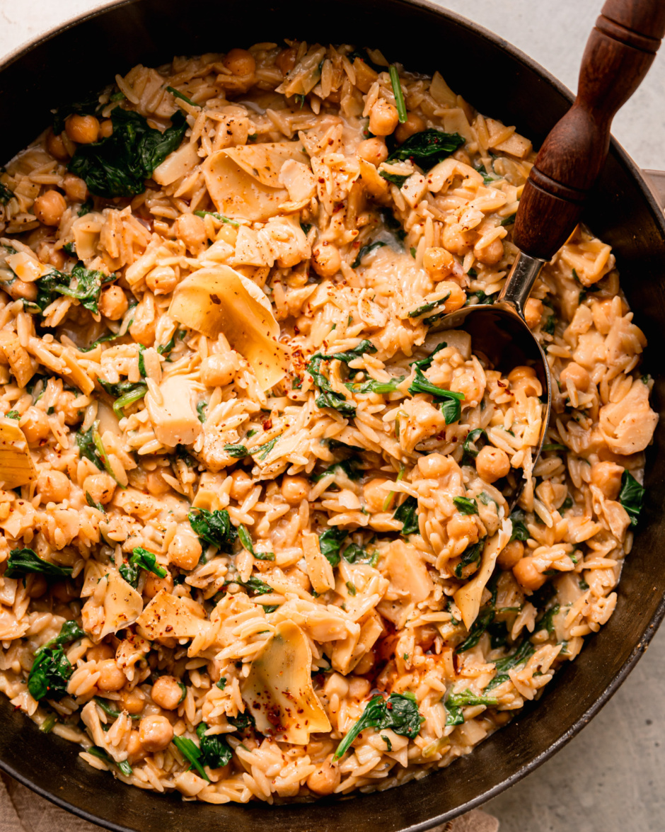 An up close, overhead shot shows a pot of vegan spinach artichoke orzo with a serving spoon sticking out.