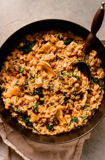 An overhead shot shows a pot of vegan spinach artichoke orzo with a serving spoon sticking out.