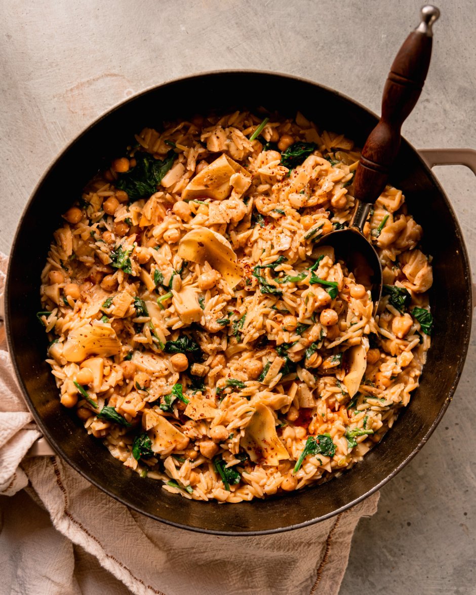 An overhead shot shows a pot of vegan spinach artichoke orzo with a serving spoon sticking out.