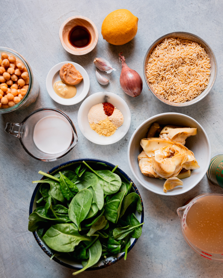 An overhead shot shows ingredients for a vegan pasta with spinach, lemon, chickpeas, artichokes, and more.