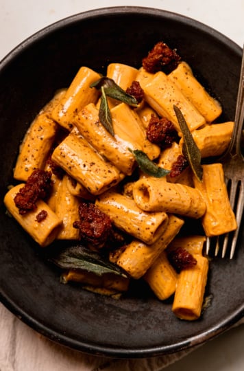 An overhead shot shows an individual serving of vegan butternut squash pasta. The sauce is very creamy and the pasta is dotted with bits of cooked veggie sausage and fried sage leaves.