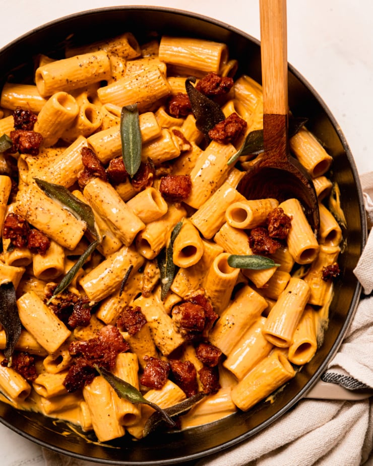 An overhead shot shows a creamy vegan butternut squash rigatoni pasta with crumbles of cooked veggie sausage and fried sage leaves. The dish is sprinkled with black pepper. A wooden serving spoon sticks out of the pot.