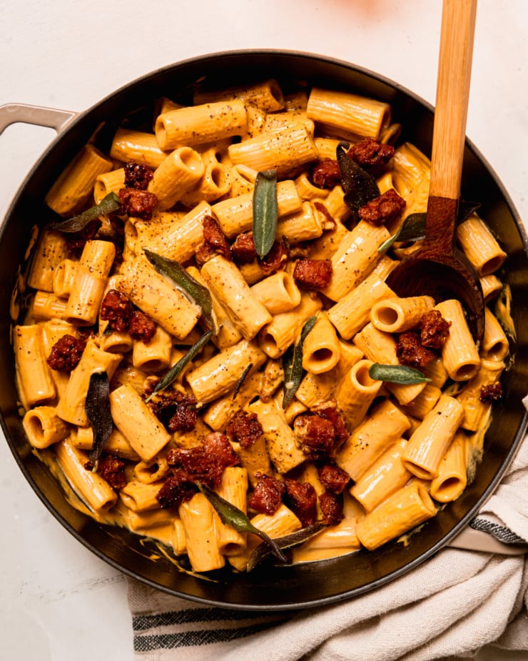 An overhead shot shows a creamy vegan butternut squash rigatoni pasta with crumbles of cooked veggie sausage and fried sage leaves. The dish is sprinkled with black pepper. A wooden serving spoon sticks out of the pot.