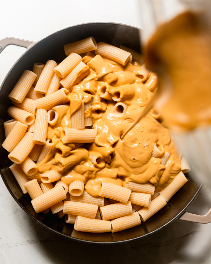 An overhead shot shows butternut squash sauce being poured over cooked rigatoni pasta in a braiser.
