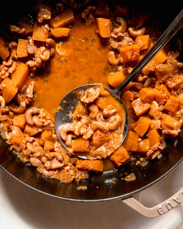 An overhead shot shows a mixture of squash, onions, cashews, walnuts, and vegetable stock in a pot with a slotted spoon.