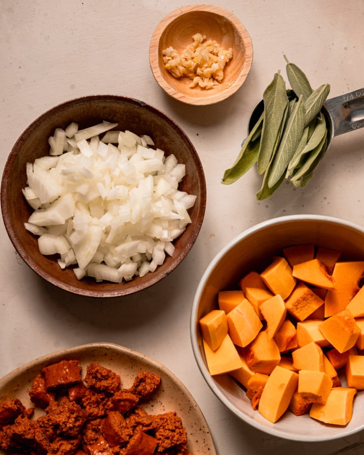 An overhead shot shows minced garlic, chopped onion, sage leaves, chopped squash, and chopped veggie sausage all in little prep bowls/plates.