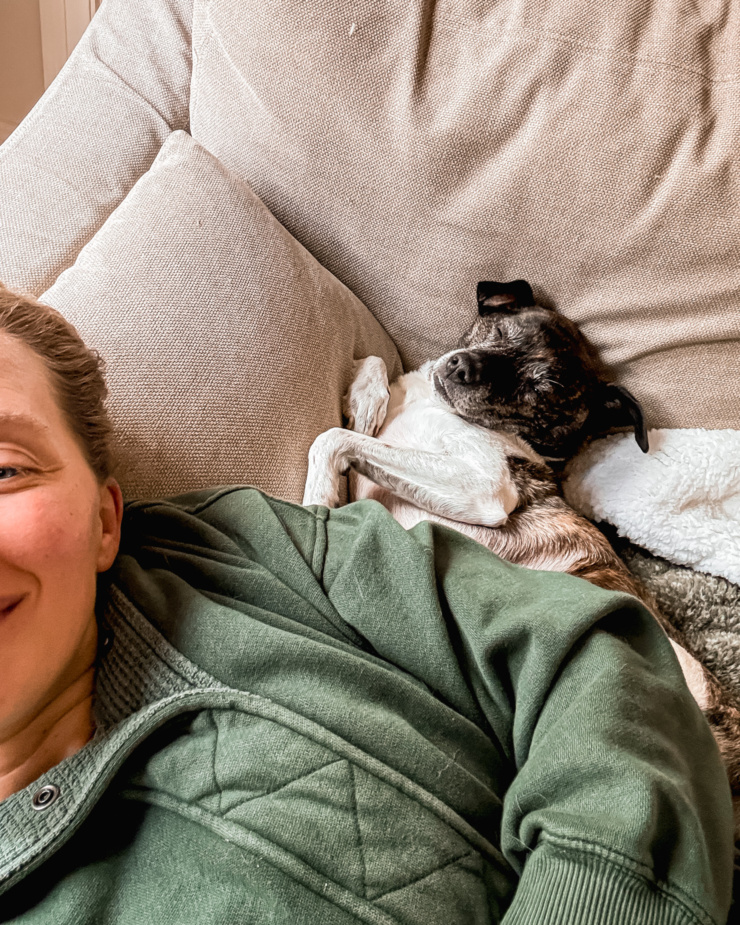 A selfie angle shows Laura and her dog Cleo laying on the couch. Cleo is wedged up on Laura next to the back cushion and is falling asleep.