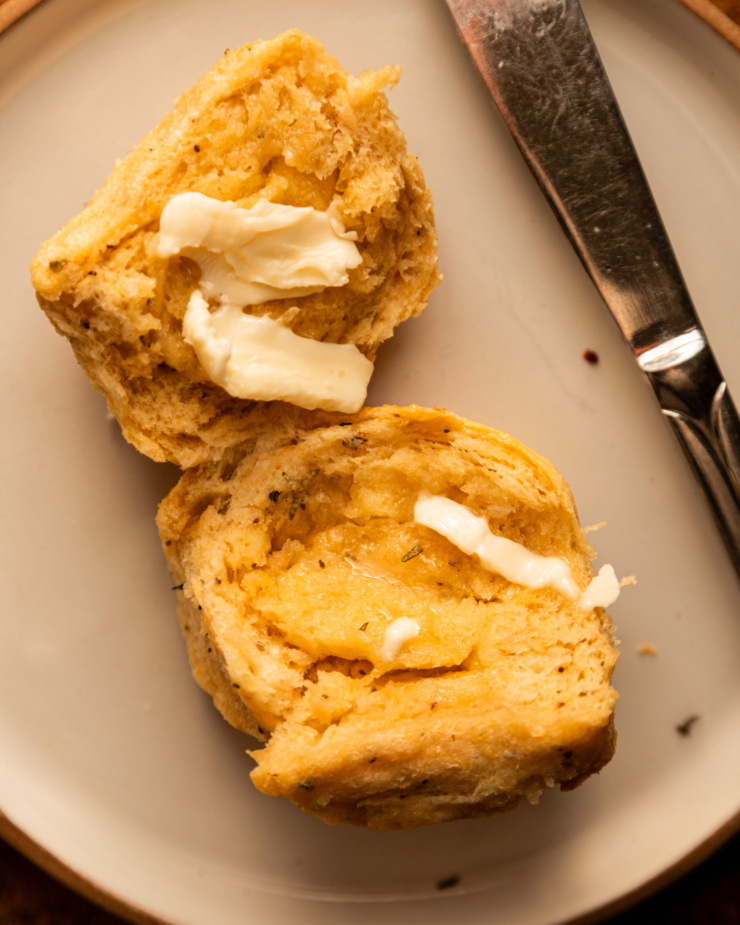 An up close, overhead shot shows a split dinner roll with vegan butter applied. The vegan butter is melting into the crevices and a butter knife is seen to the side.