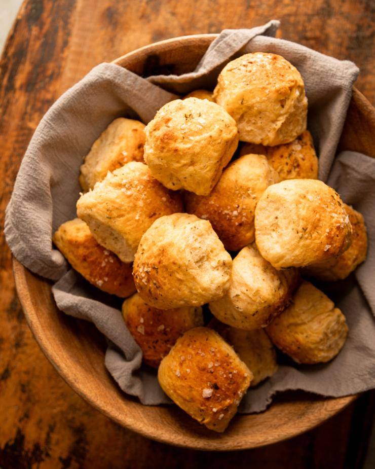 An overhead shot shows a bowl lined with linen and filled with no-knead dinner rolls.