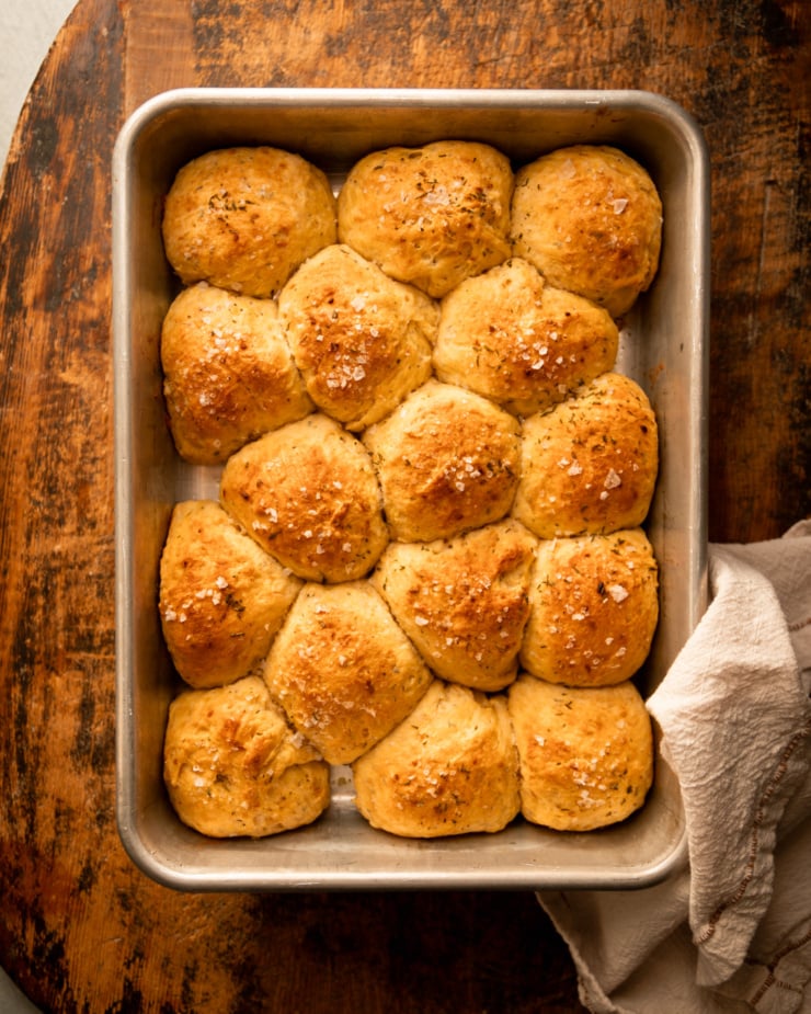 An overhead shot shows a baking dish filled with no-knead dinner rolls that feature sweet potato purée and chopped herbs. The rolls are topped with flaky salt.