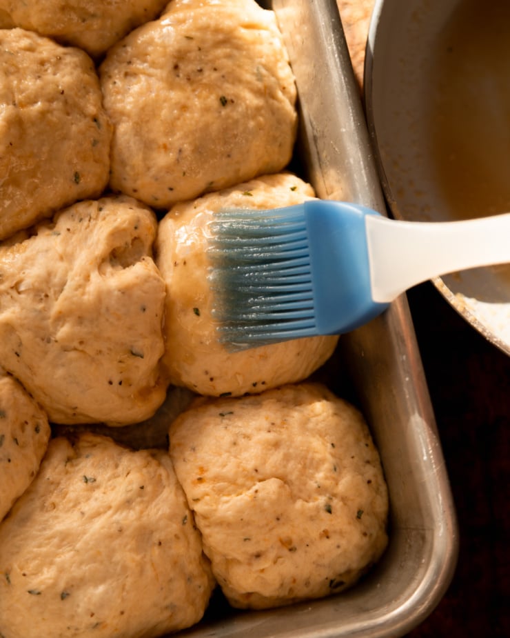 An up close, overhead shot shows a brush applying melted vegan butter to proofed balls of dough in a baking dish.