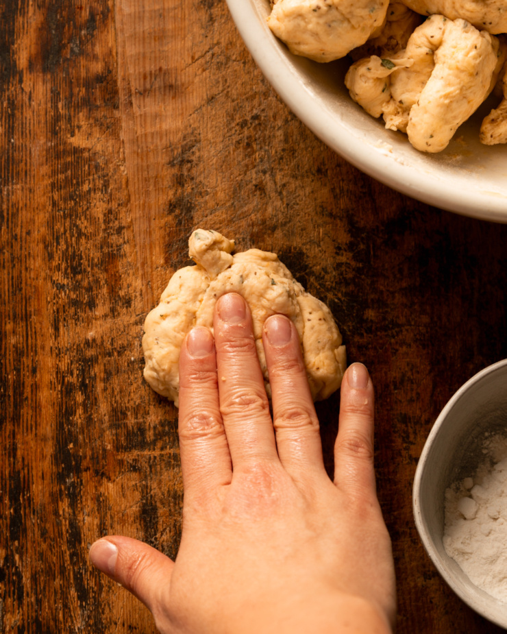 An overhead shot shows a hand pressing down on a portion of dough on a wooden board.