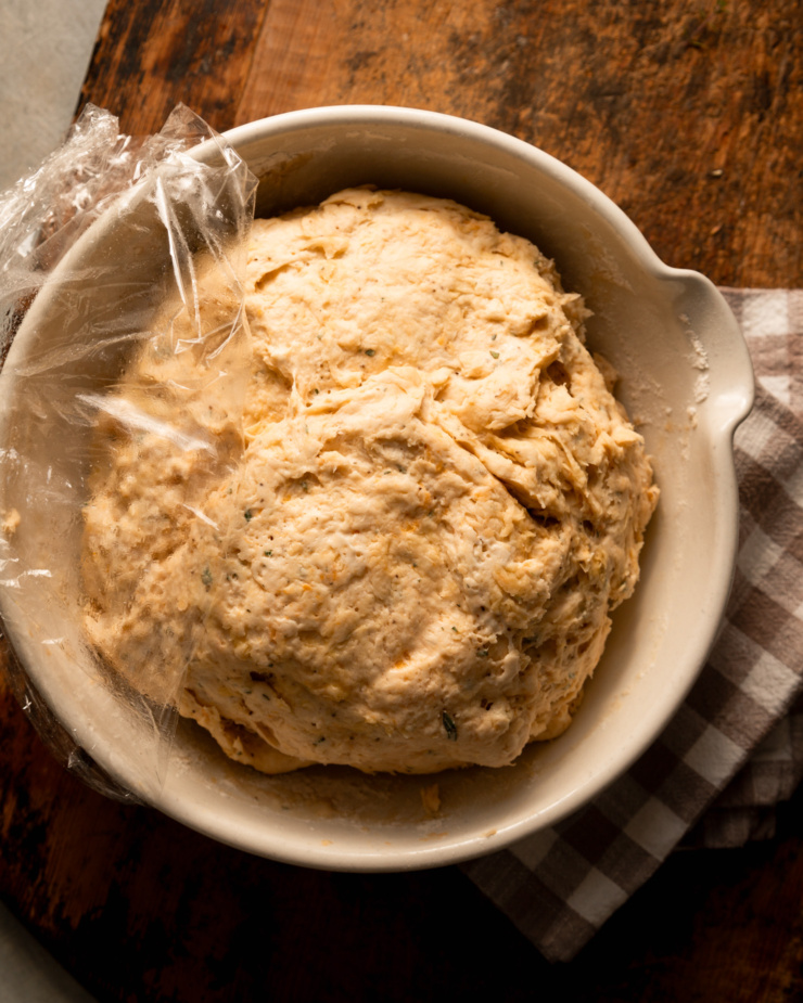 An overhead shot shows a proofed bread dough, flecked with spices and herbs, in a large bowl.