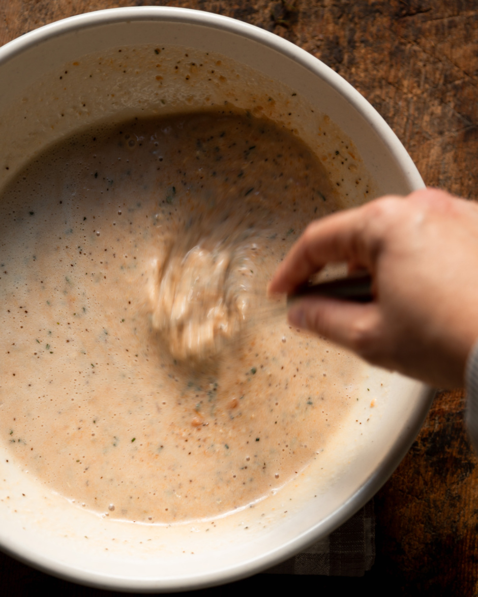 An overhead shot shows a hand using a whisk to bring together an herb-flecked milk mixture in a bowl.