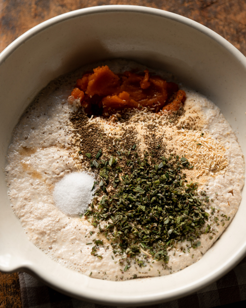 An overhead shot shows a bowl with soy milk, yeast, salt, chopped herbs, sweet potato purée, celery seed, garlic powder, and dehydrated minced onion.