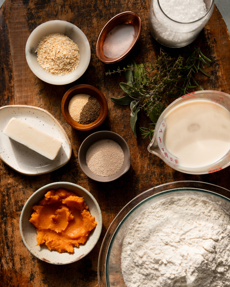 An overhead shot shows ingredients for vegan no-knead dinner rolls: flaky salt, fine salt, sage, rosemary, thyme, soy milk, active dry yeast, all purpose flour, sweet potato purée, vegan butter, garlic powder, celery seed, and dehydrated minced onion.