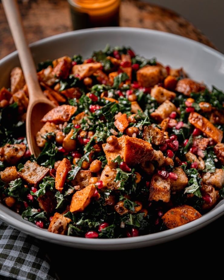 An up close, 3/4 angle shot shows a Thanksgiving Panzanella salad, containing kale, roasted sweet potato, carrot, parsnip, toasted bread cubes, and pomegranate seeds. A wooden spoon is sticking out of the salad.
