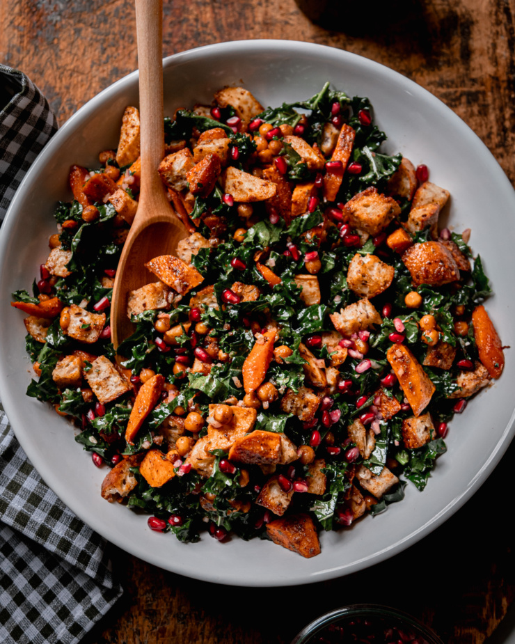 An overhead shot shows a Thanksgiving Panzanella salad, containing kale, roasted sweet potato, carrot, parsnip, toasted bread cubes, and pomegranate seeds. A wooden spoon is sticking out of the salad.