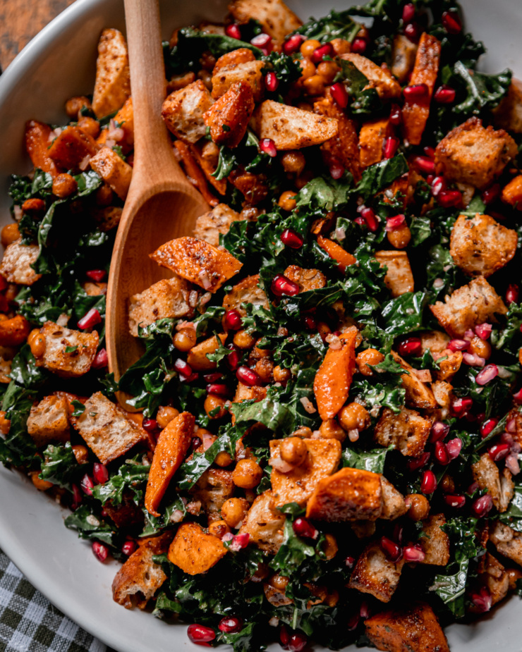 An up close, overhead shot shows a Thanksgiving Panzanella salad, containing kale, roasted sweet potato, carrot, parsnip, toasted bread cubes, and pomegranate seeds. A wooden spoon is sticking out of the salad.