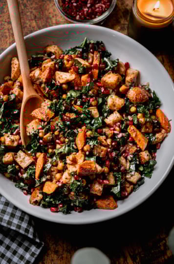 An overhead shot shows a Thanksgiving Panzanella salad, containing kale, roasted sweet potato, carrot, parsnip, toasted bread cubes, and pomegranate seeds. A lit candle is nearby as well as a bowl of more pomegranate seeds. A wooden spoon is sticking out of the salad.