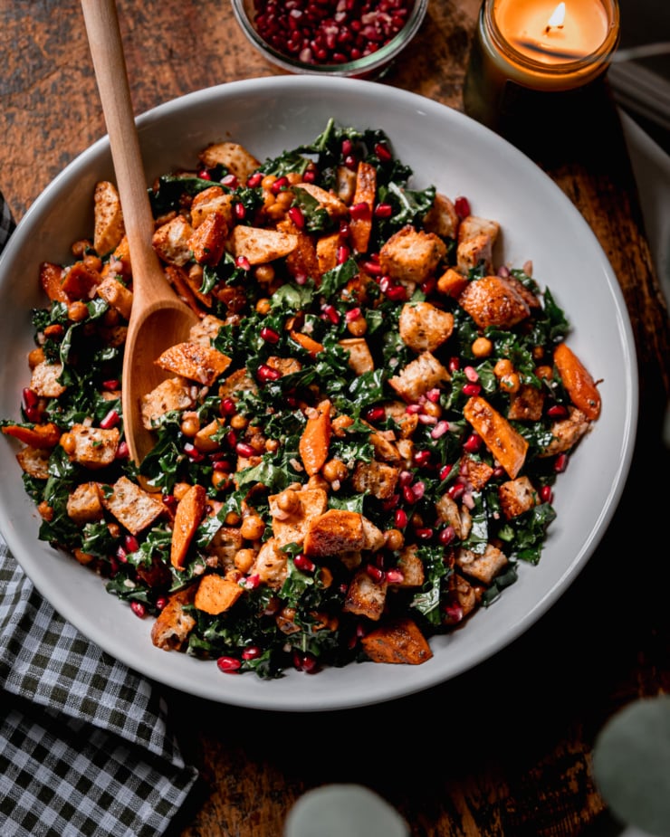 An overhead shot shows a Thanksgiving Panzanella salad, containing kale, roasted sweet potato, carrot, parsnip, toasted bread cubes, and pomegranate seeds. A lit candle is nearby as well as a bowl of more pomegranate seeds. A wooden spoon is sticking out of the salad.