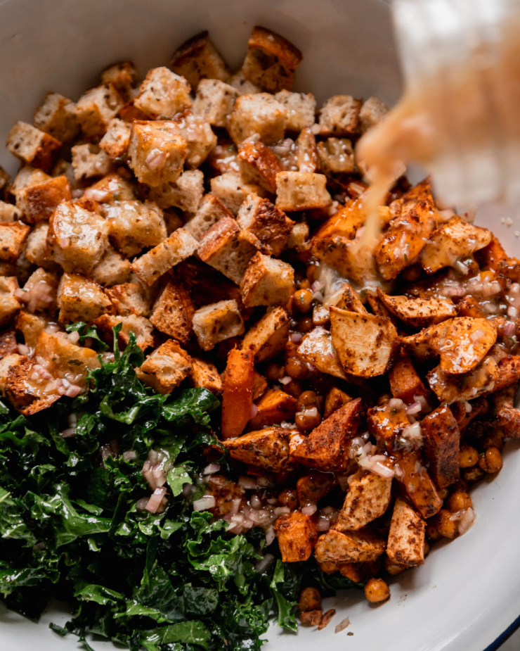 An overhead shot shows salad dressing being poured over toasted bread cubes, chopped kale, roasted vegetables, and chickpeas.