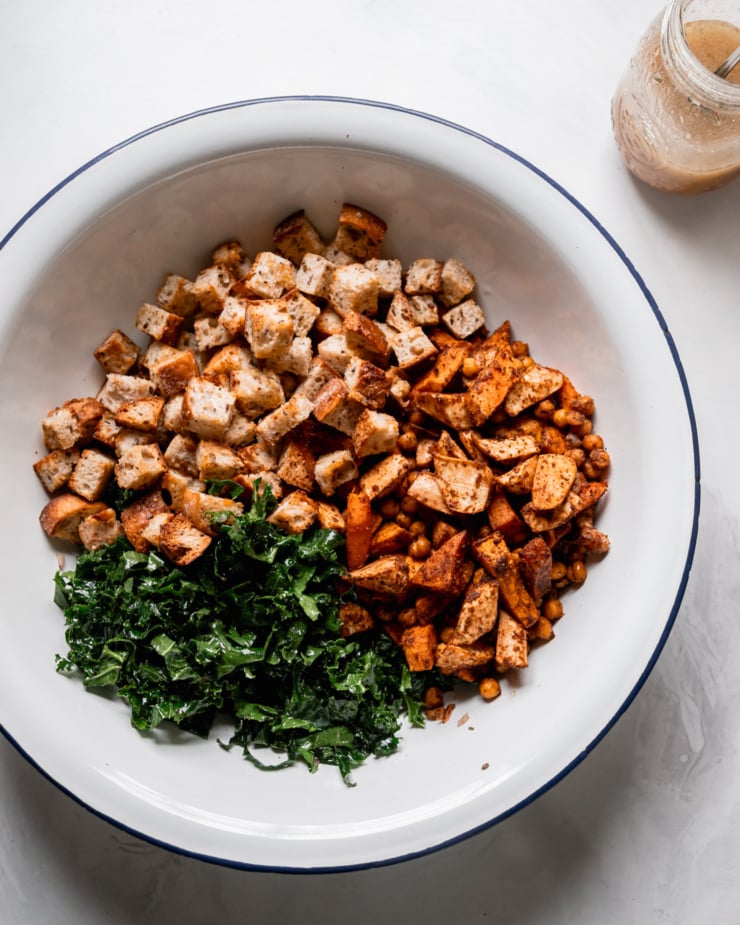 An overhead shot shows a large bowl, containing toasted bread cubes, chopped and dressed kale, and a mix of roasted sweet potato, parsnip, carrot, and chickpeas.
