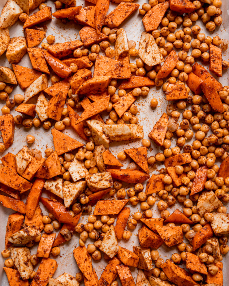 An overhead shot shows chickpeas, chopped sweet potato, carrot, and parsnip all coated in oil and spices on a baking sheet.