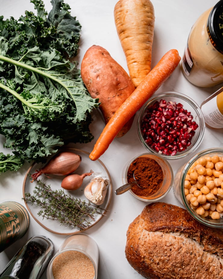 An overhead shot shows ingredients for a salad: parsnip, carrot, sweet potato, dijon, pomegranate seeds, spices, chickpeas, multigrain bread, thyme, shallots, garlic, olive oil, vinegar, and kale.