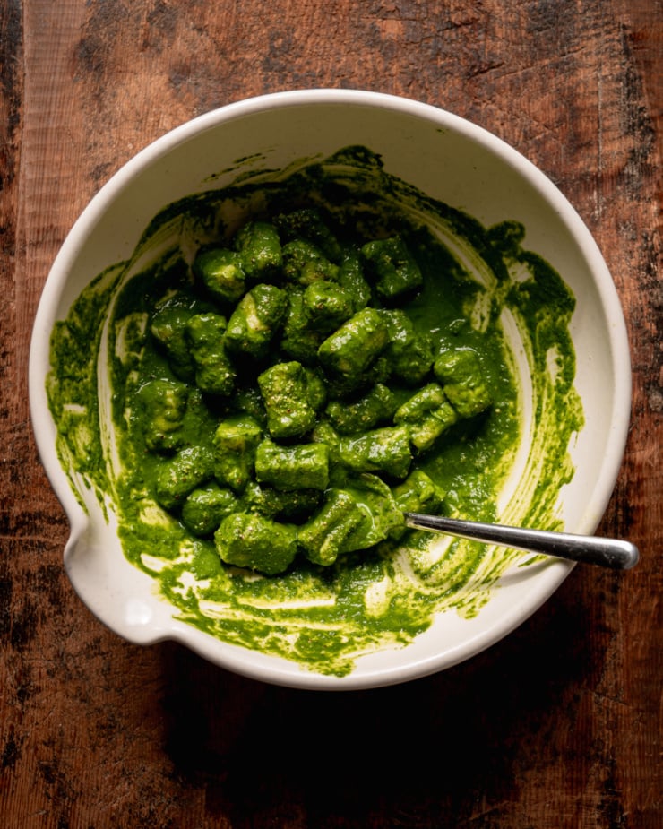 An overhead shot shows homemade gnocchi tossed in a deep green sauce in a bowl. There is a spoon sticking out of the bowl.