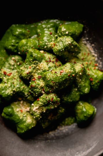 An up close, overhead shot shows a serving of homemade vegan gnocchi covered in a thick green kale sauce. The dish is garnished with vegan "parmesan" and Aleppo chili
