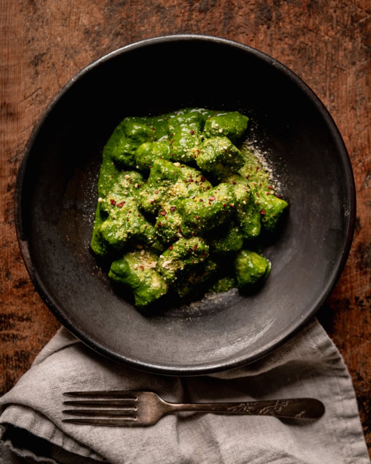 An overhead shot shows a serving of homemade vegan gnocchi covered in a thick green kale sauce. The dish is garnished with vegan "parmesan" and Aleppo chili. A linen napkin and fork are nearby.