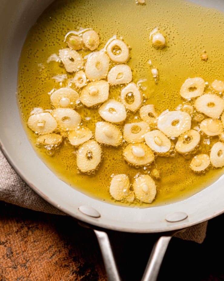 An overhead shot shows sliced garlic sizzling in olive oil in a small skillet.
