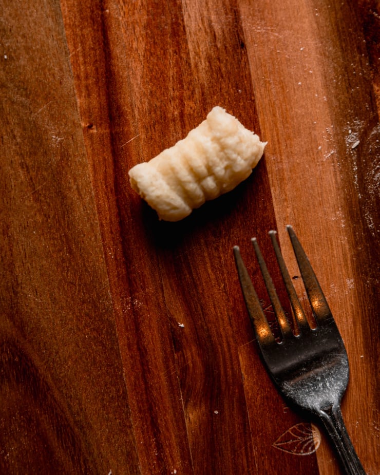 An overhead shot shows a singular gnocchi with fork tine indentations on it next to a fork.