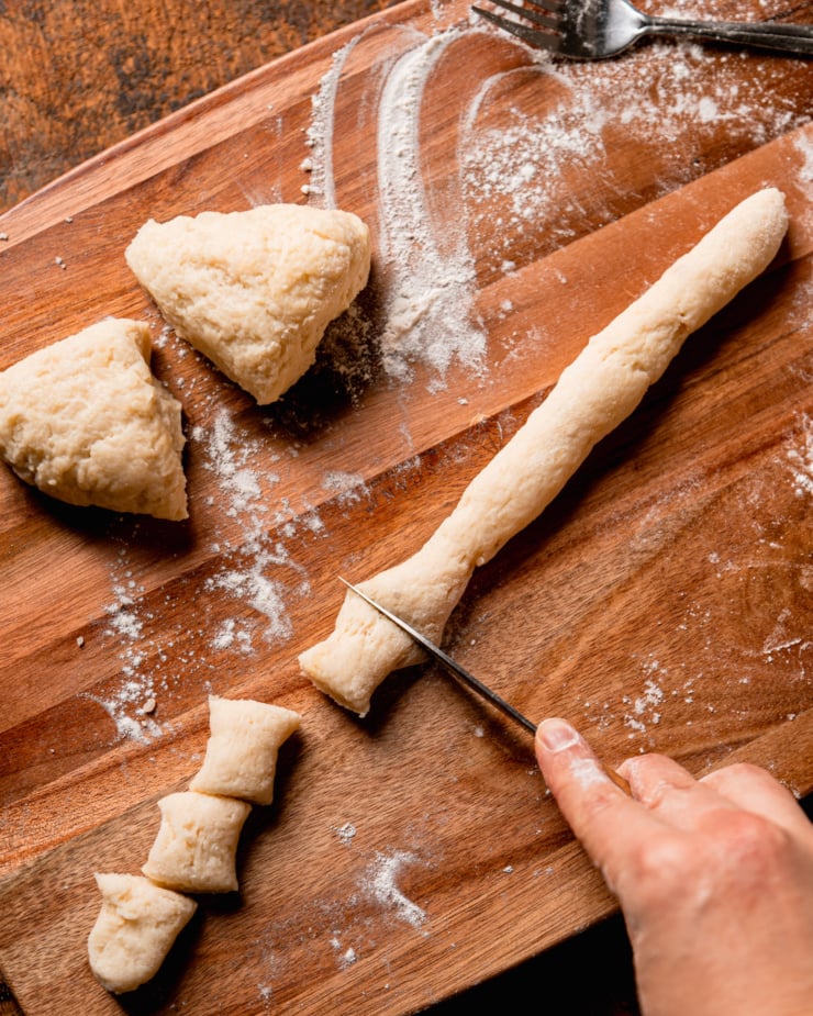 An overhead shot shows a hand using a knife to cut portions of dough on a wooden cutting board.