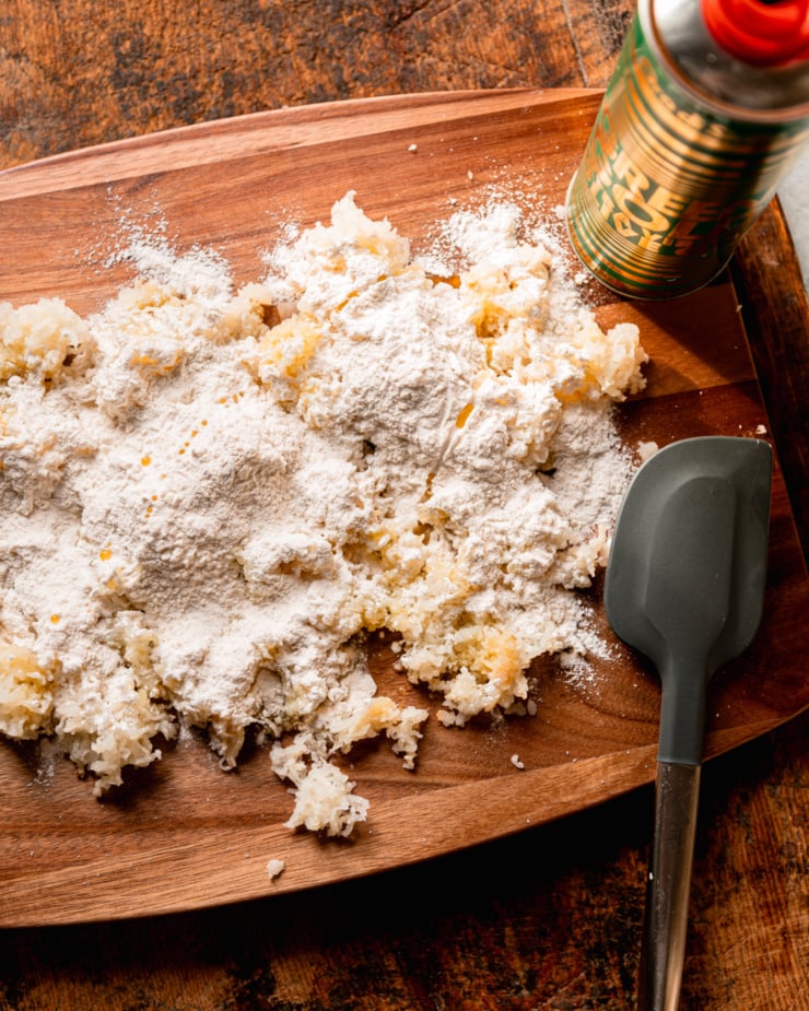 An overhead shot shows a cutting board covered in finely grated cooked potato, flour, and olive oil. A rubber spatula is nearby.