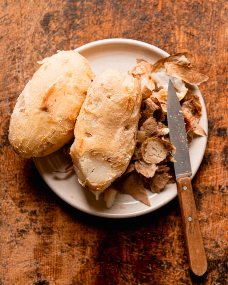 An overhead shot shows two peeled, baked russet potatoes on a plate, along with the discarded peels and a paring knife.