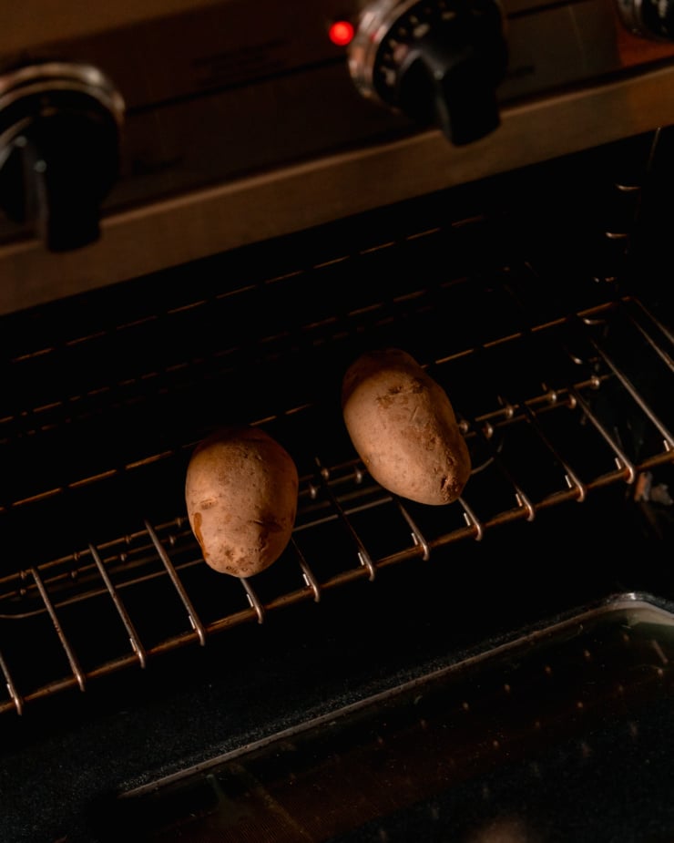 An overhead shot shows two russet potatoes on an oven rack, about to be baked.